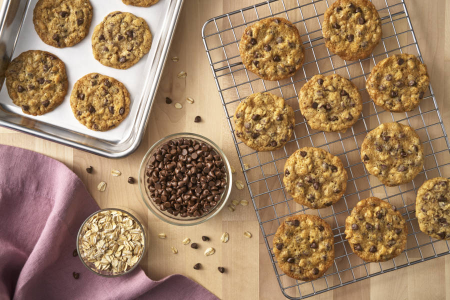 Photograph of oatmeal chocolate-chip cookies on cooling rack and pan, bowls of oats and chips